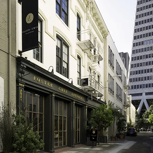 Exterior of Foster Gwin Antiques storefront on a city street with tall buildings and greenery.