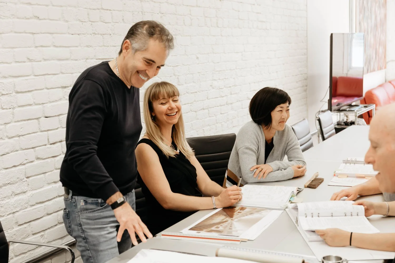 Four people in a meeting room with white brick wall, smiling and talking at a table with open books and documents. One man stands, the others sit, in a casual office setting.