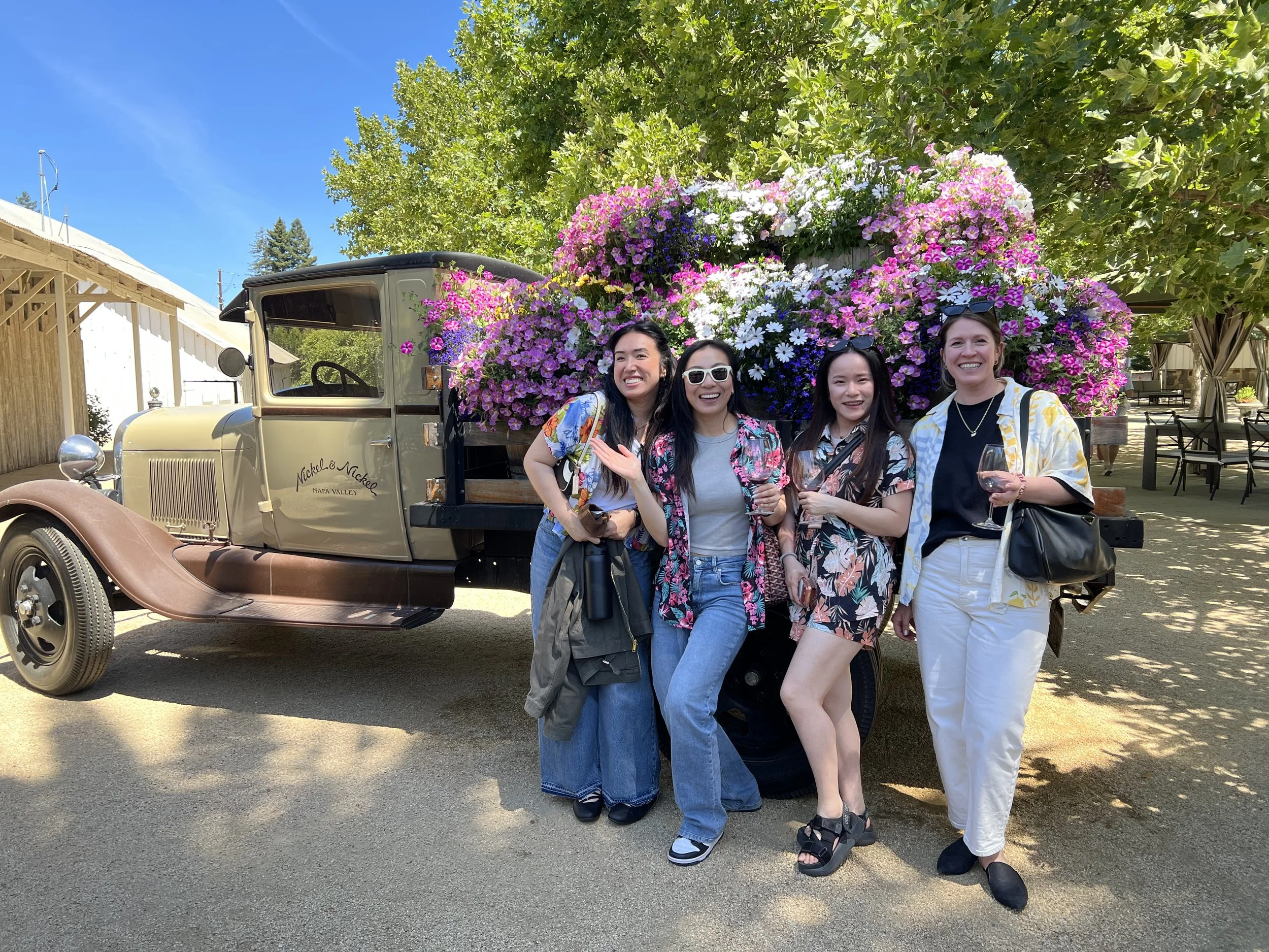 Four women standing in front of a vintage truck filled with pink, white, and purple flowers, smiling and holding glasses of wine on a sunny day.