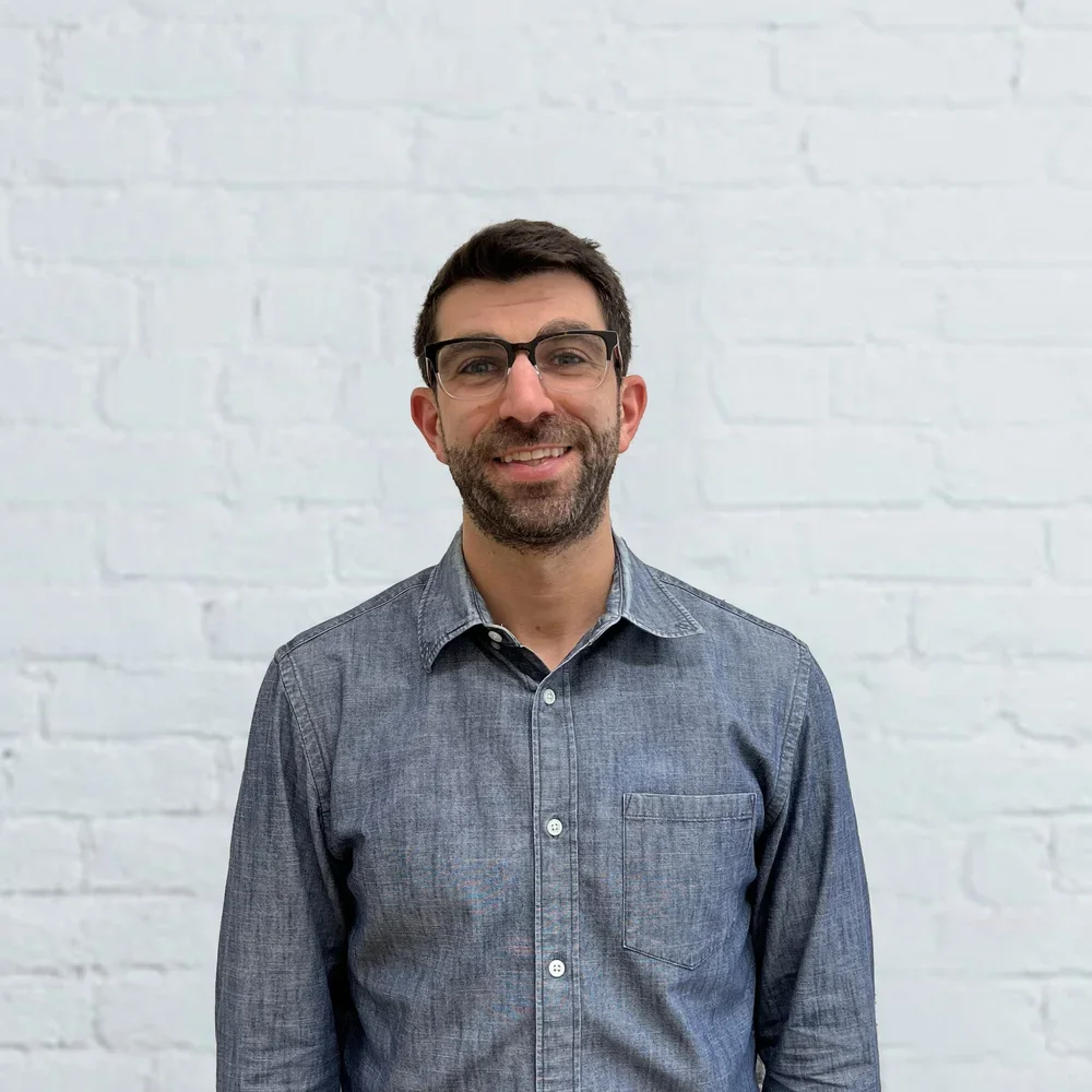 A man with glasses and short brown hair, wearing a gray button-up shirt, smiling and standing in front of a white brick wall.