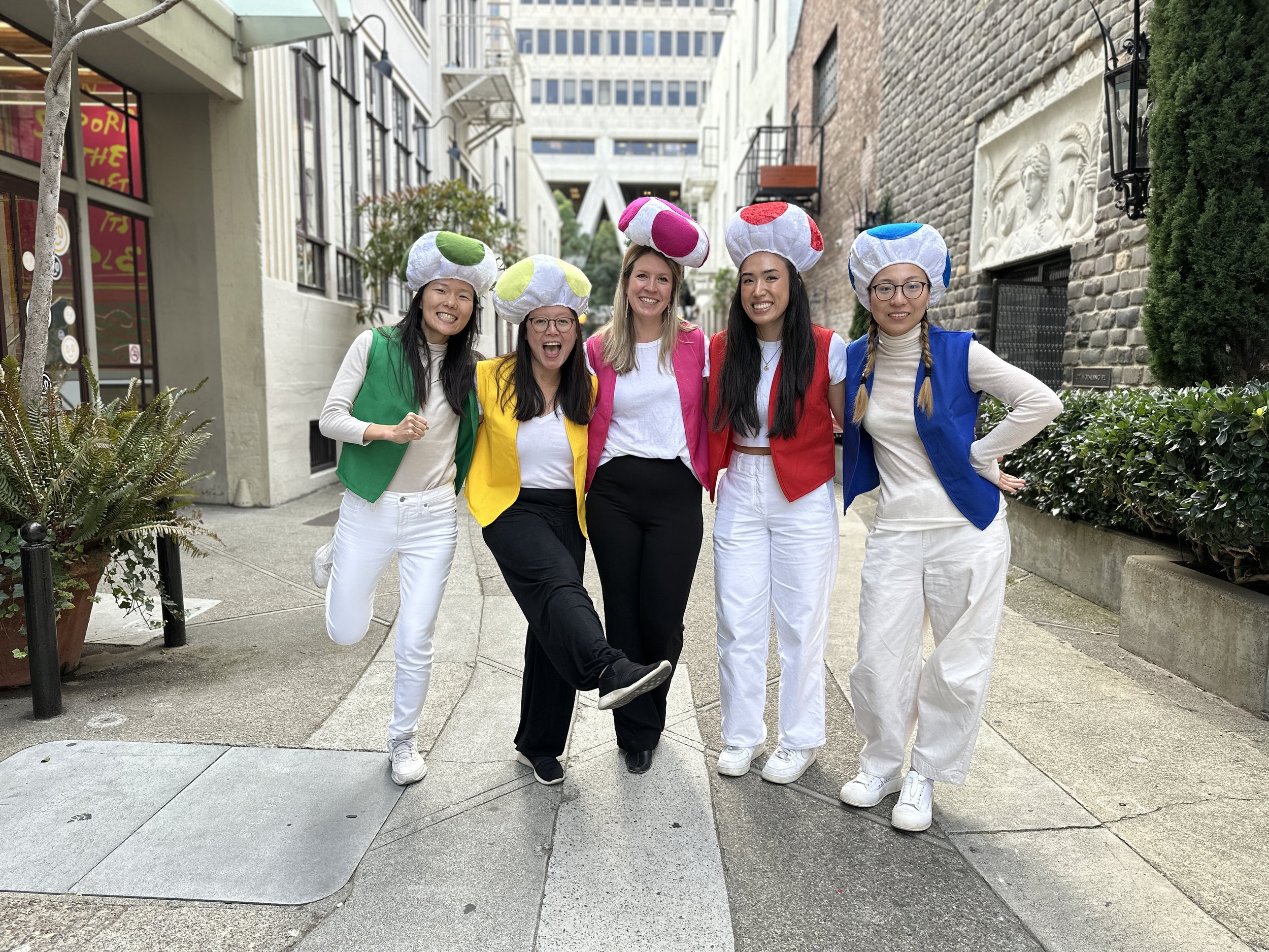 Five women wearing mushroom-shaped hats in various colors are standing close together on a city sidewalk, smiling and posing for a photo. They are dressed in casual clothing with colorful vests, and one woman is lifting her leg playfully. The backgro