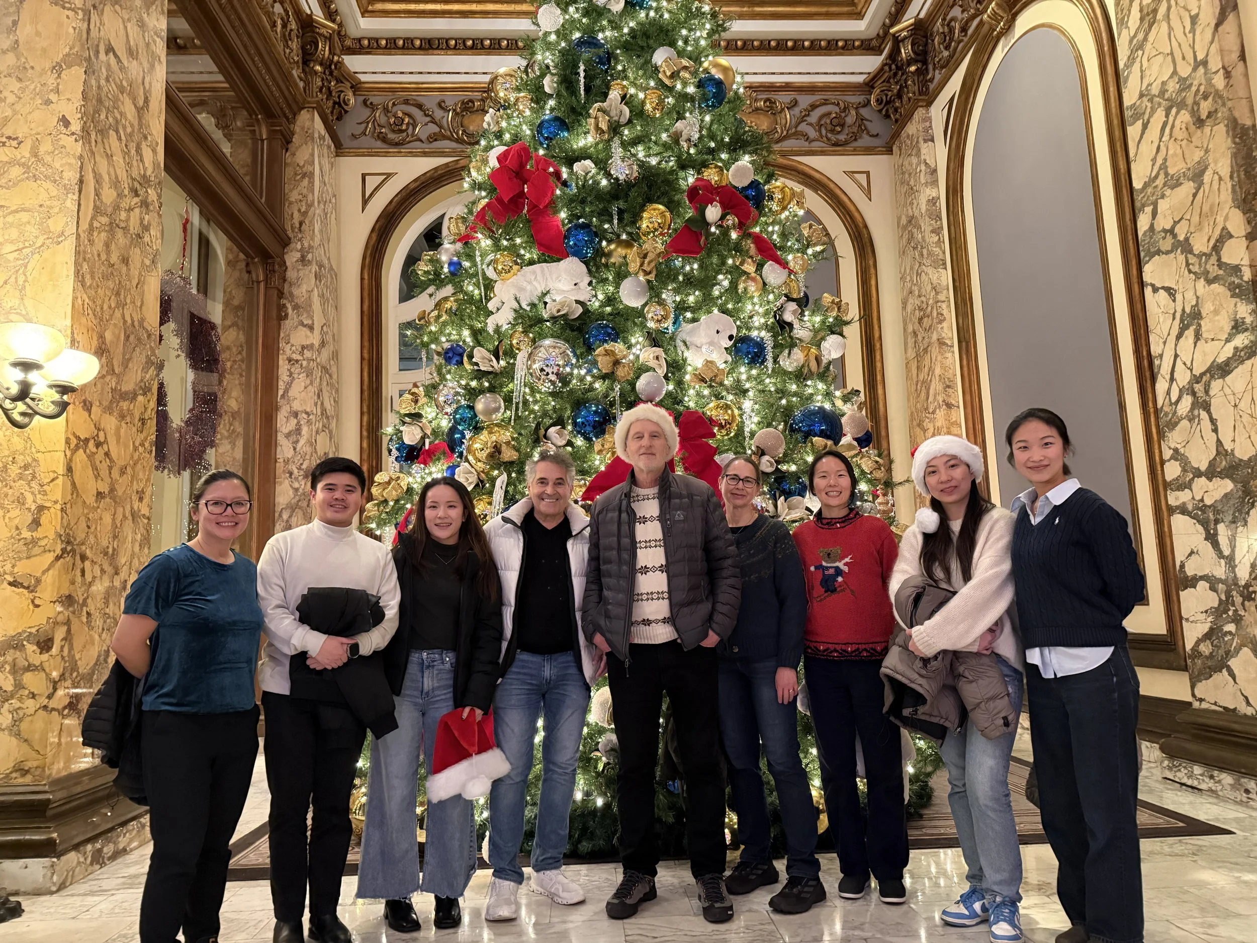 A group of nine people standing in front of a large decorated Christmas tree in a luxurious, ornate room with marble walls and gold accents. The group includes men and women, some wearing Santa hats and Christmas sweaters, smiling for the photo.