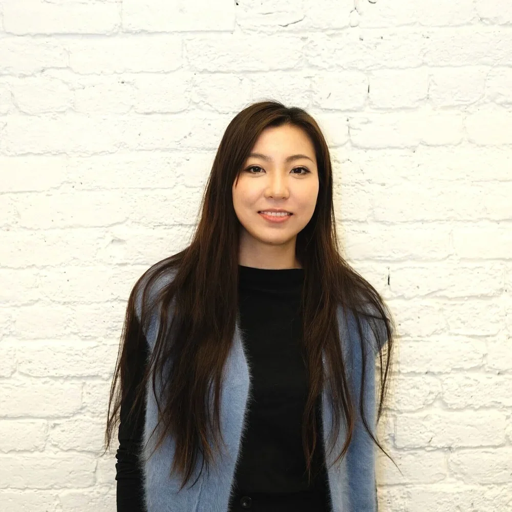A young woman with long dark hair, wearing a black top and a light blue vest, standing against a white brick wall and smiling at the camera.