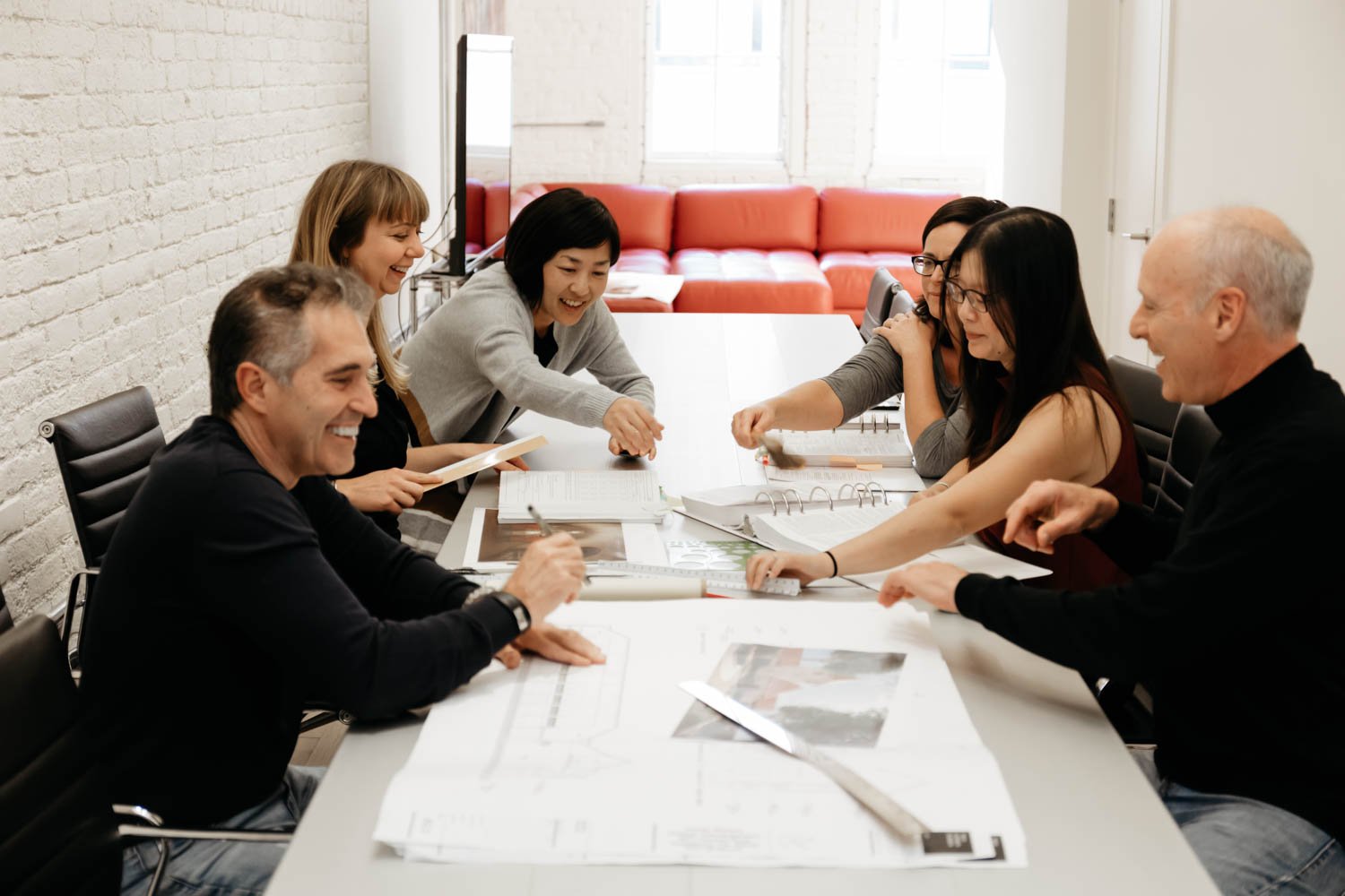 Six people smiling and discussing around a white table in a bright office room, with documents, books, and a ruler on the table.