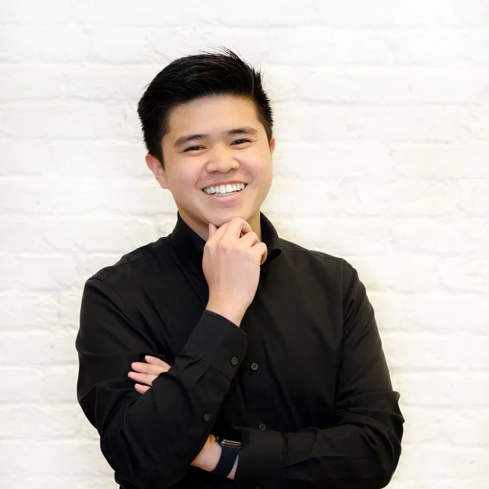 A young man with short black hair, wearing a black shirt, smiling, standing against a white brick wall.
