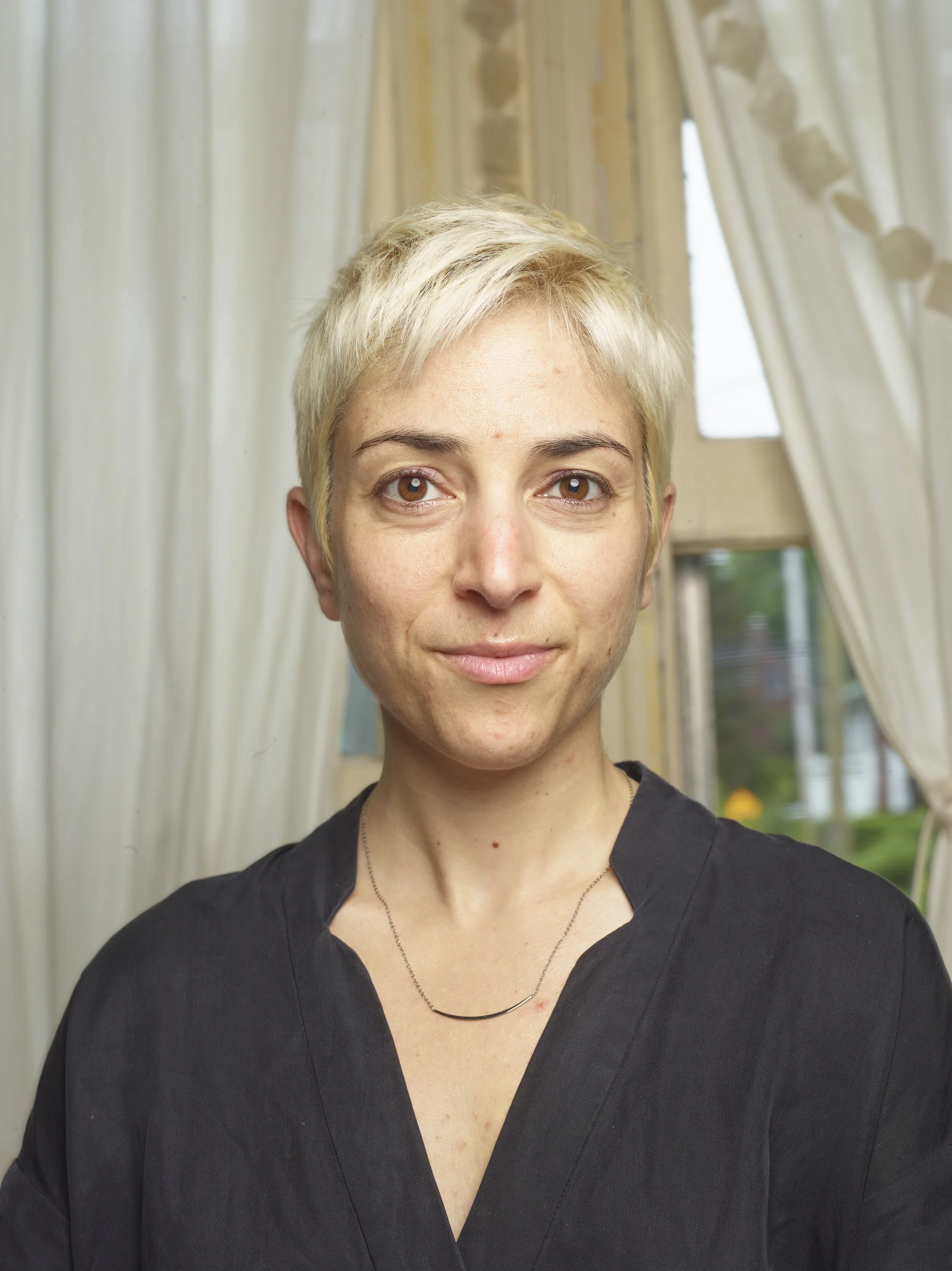 Headshot of a woman with short blonde hair, wearing a dark grey top and small necklace, standing in a room with light-colored curtains and windows in the background.