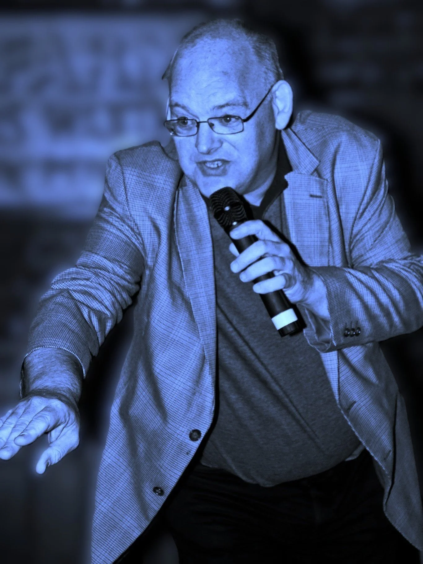 A man wearing glasses, a checkered blazer, and a dark shirt, speaking into a microphone, with a blurred background.