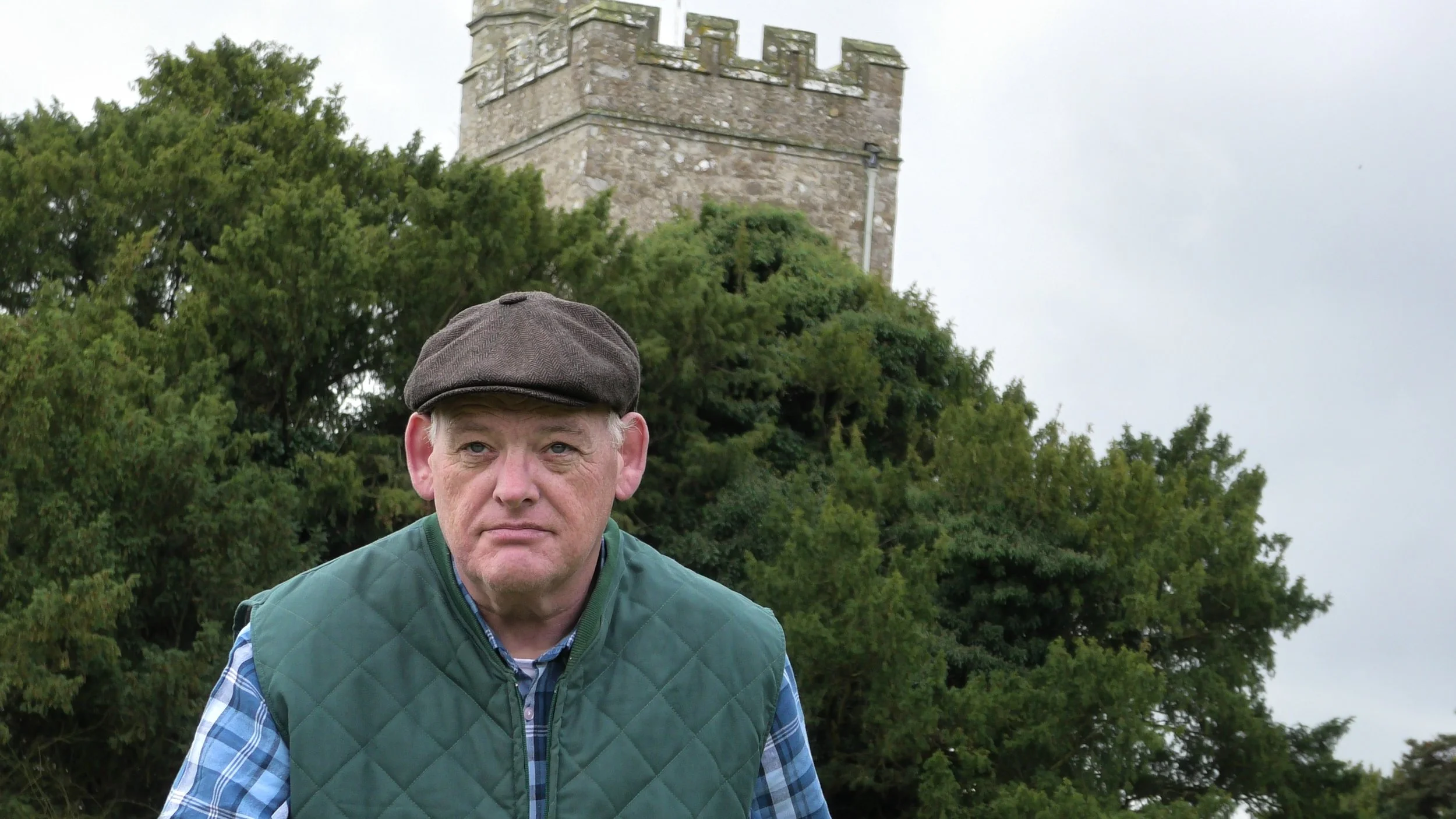 A man wearing a brown flat cap, a blue and white checkered shirt, and a green quilted vest standing outdoors with trees and a stone tower in the background.