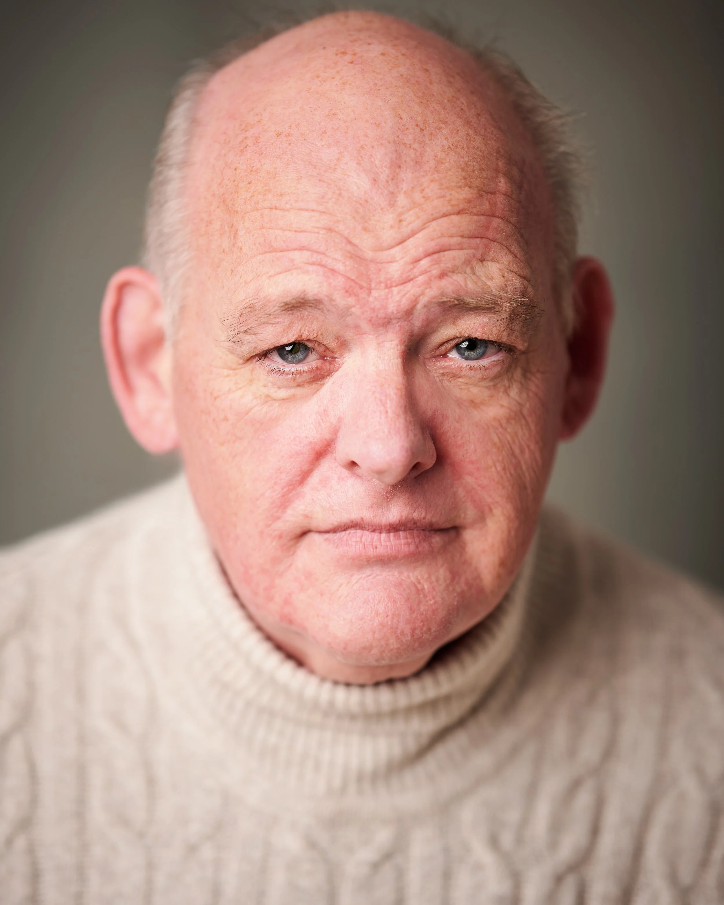 Close-up of an elderly person's face, showing a bald head, light skin with freckles, and wearing a beige knitted turtleneck sweater.