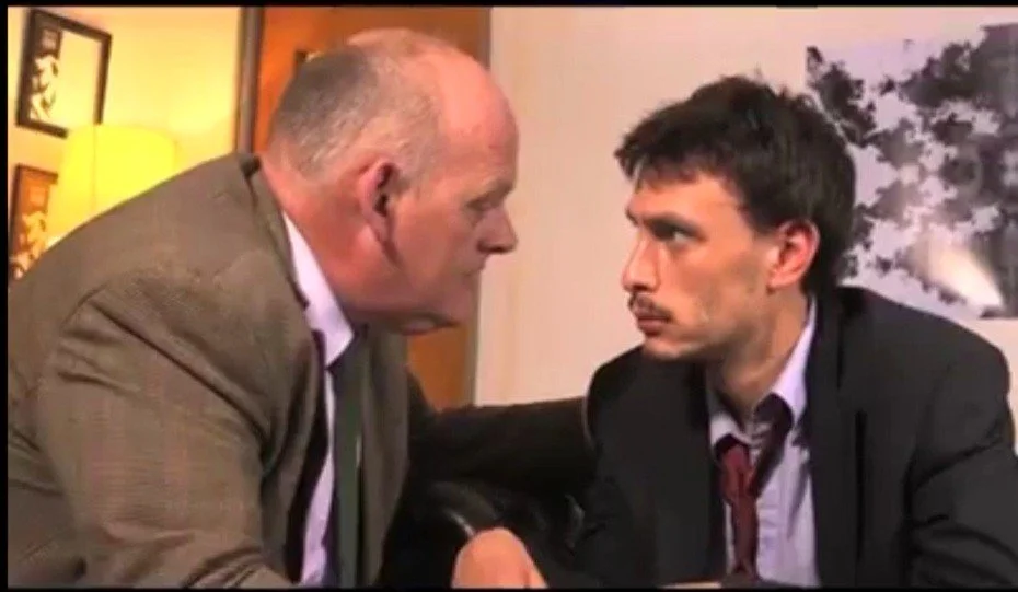 Two men are facing each other very closely inside a room, having an intense conversation. The older man on the left has a bald head with gray hair on the sides and is wearing a brown blazer. The younger man on the right has dark hair, a short beard, and is dressed in a black suit with a white shirt and a loosened maroon tie.
