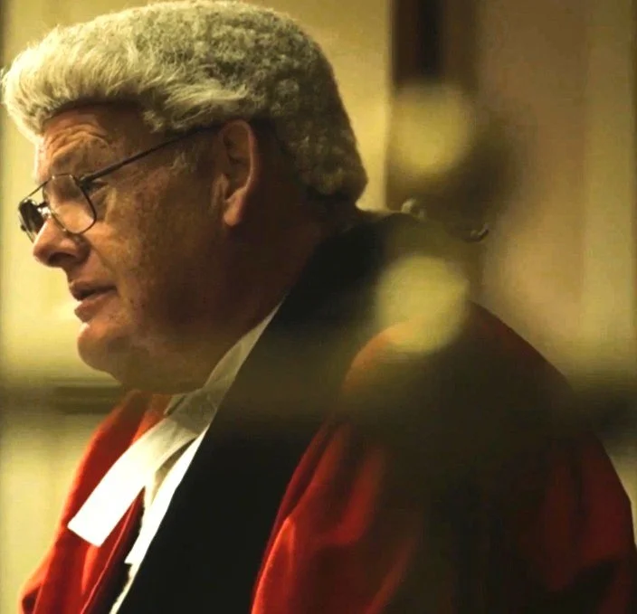 Profile of an older man with gray hair, wearing glasses and a red jacket, in a dimly lit indoor setting.