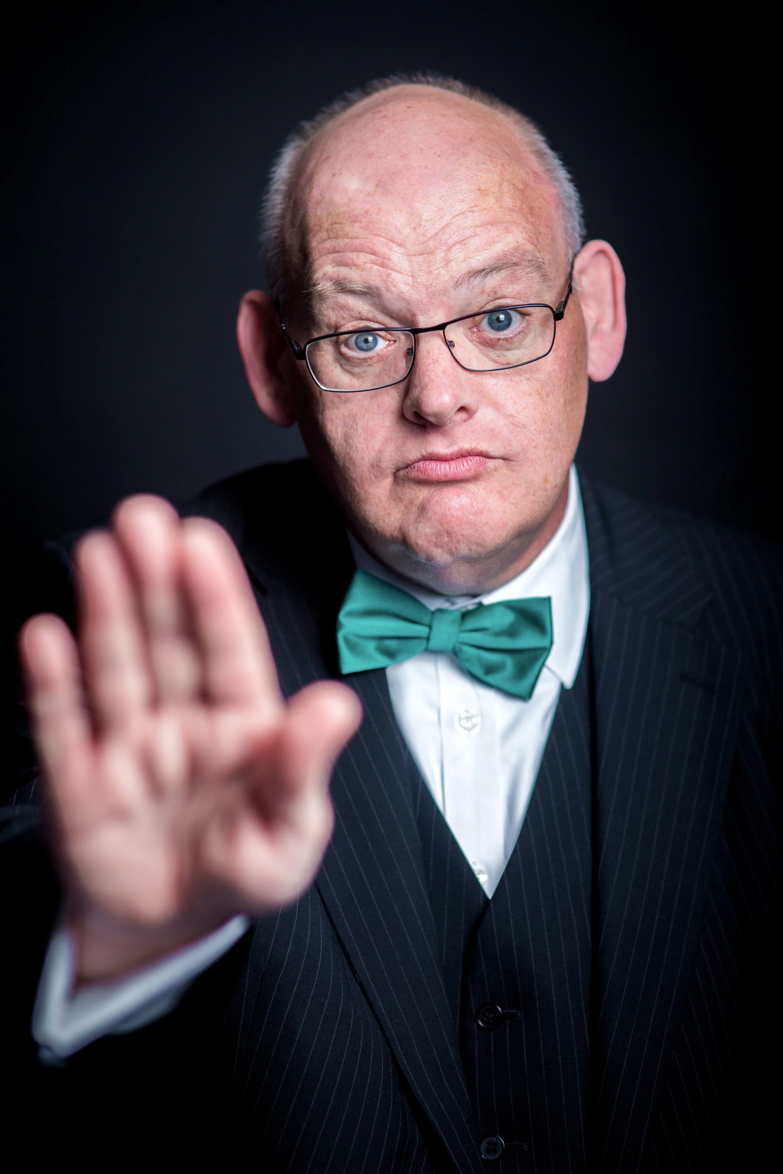 A man with glasses, light skin, and wearing a tuxedo with a green bow tie, reaching out with his hand towards the camera, with a dark background.