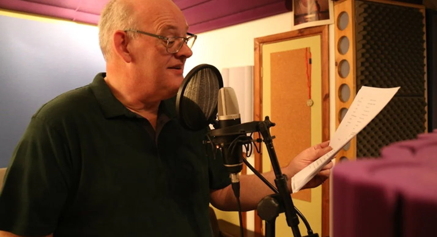 An elderly man with glasses in a recording studio, standing in front of a microphone, reading from a script or lyrics.