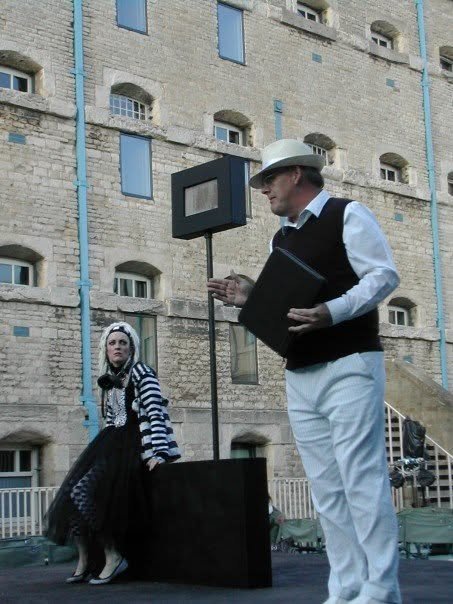 A man in a white suit and hat stands outdoors in front of a brick building, holding a black folder and gesturing towards a rectangular black device on a pole. A woman in a striped shirt and black skirt stands nearby, looking at him.