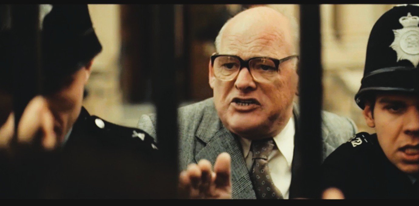 An older man with glasses and a suit is speaking to two police officers behind bars.