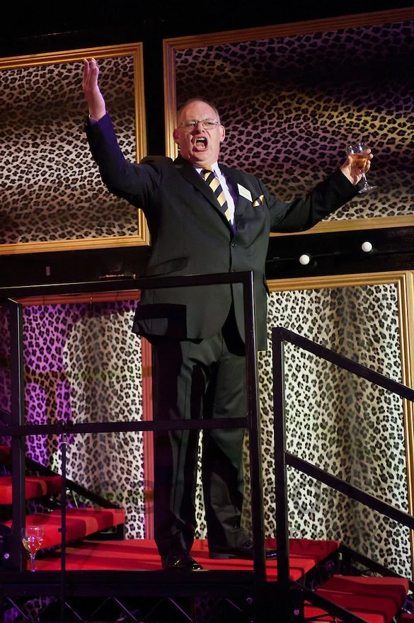 A man in a suit on a stage with leopard print background, raising one arm and holding a drink, appearing to make a toast or announcement.