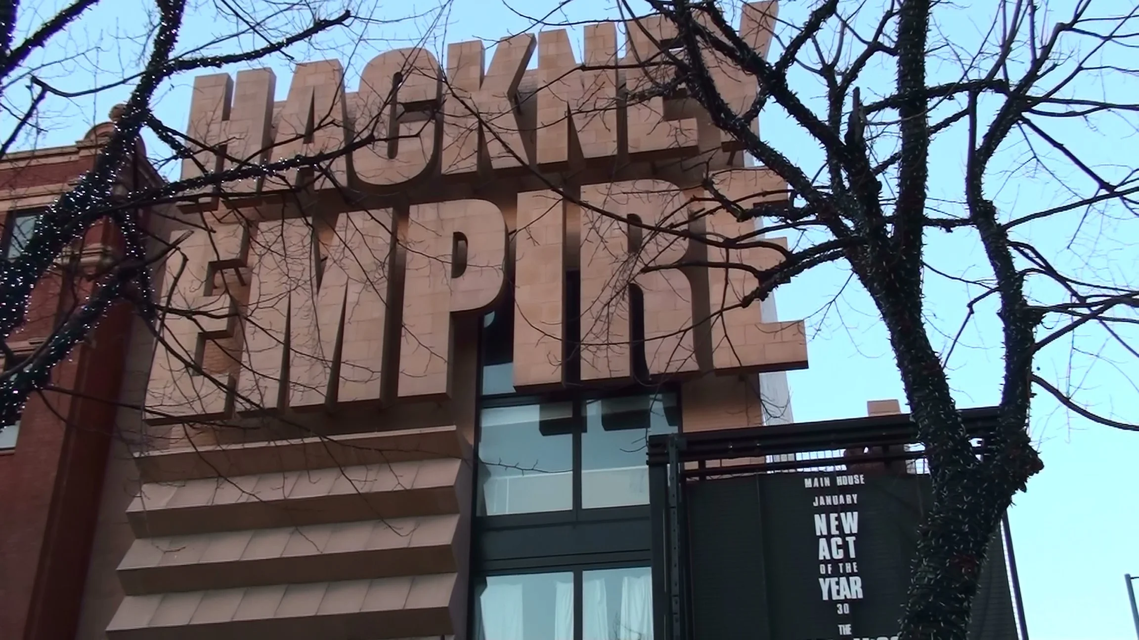 View of a modern building with large 3D letters spelling 'WHEEL' on the facade, partially obscured by leafless trees with string lights, and a digital sign displaying text in the foreground.