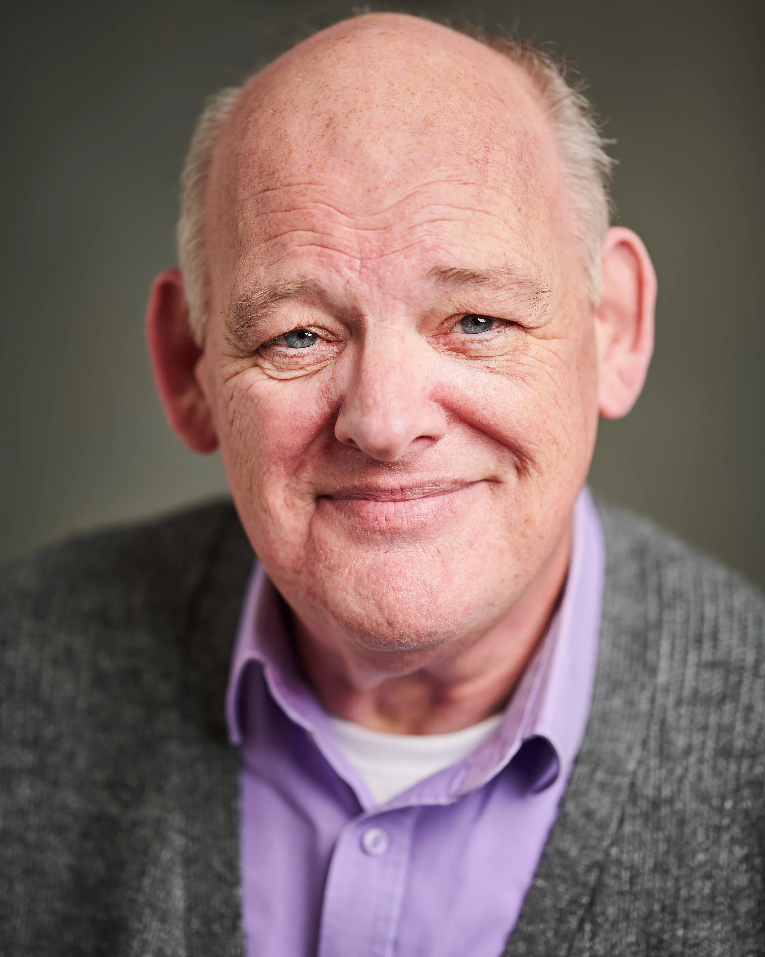 Close-up of an older man with blue eyes, receding hairline, wearing a purple shirt and gray sweater, smiling slightly, with a dark background.