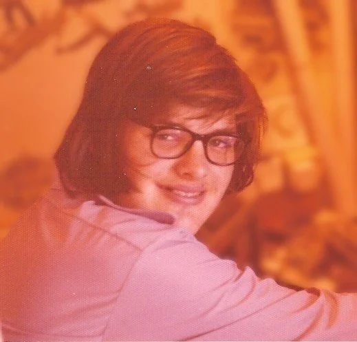 A person with shoulder-length brown hair, glasses, and a light pink shirt, smiling and looking over their shoulder.