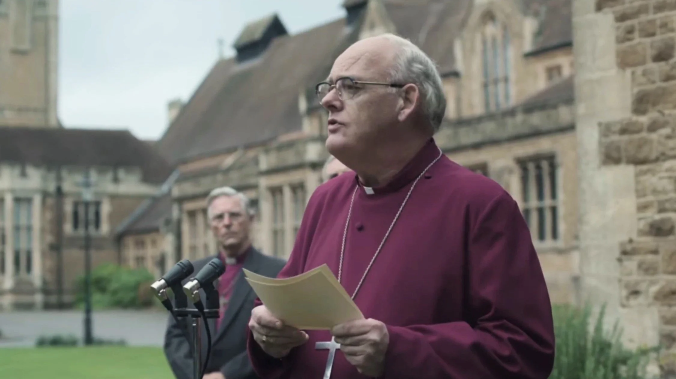 A clergyman in a red robe and glasses speaking at a podium with microphones outdoors, with historic stone buildings in the background.