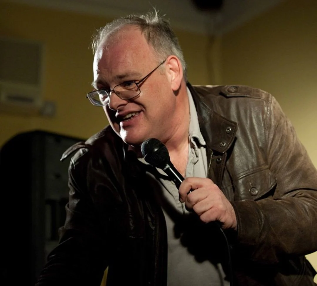 A middle-aged man with glasses and short hair, wearing a brown leather jacket, is holding a microphone and smiling during a performance or speech, with a yellow wall background.