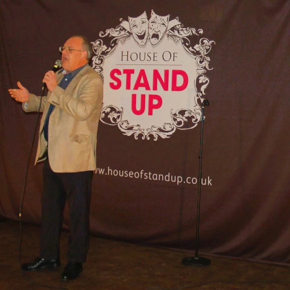 A man in glasses, a beige blazer, and dark pants speaking into a microphone on stage, with a backdrop that reads 'House of Stand Up' and features comedy masks.