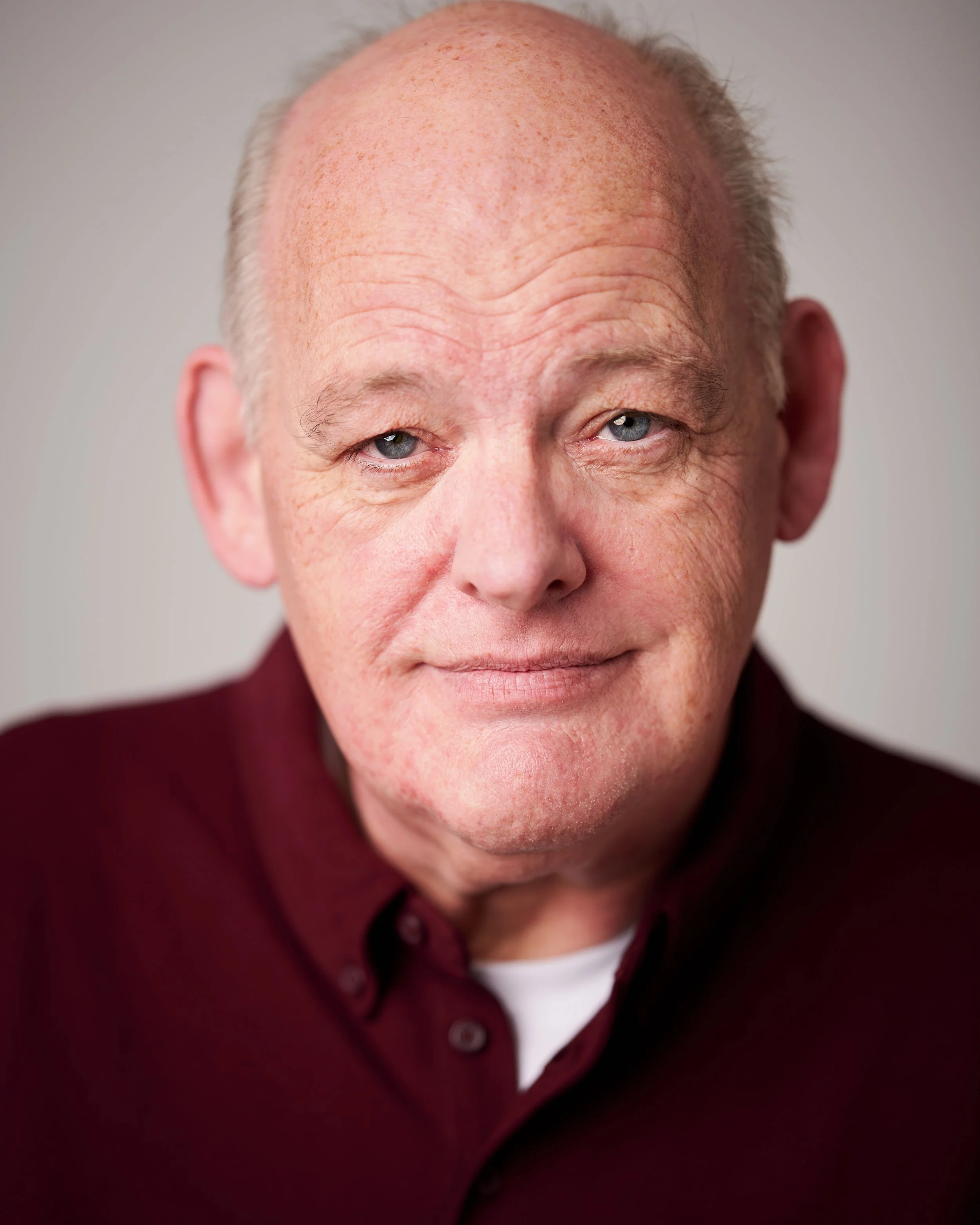 Close-up of an elderly man with a bald head, blue eyes, and faint wrinkles, wearing a dark red shirt and a white undershirt.
