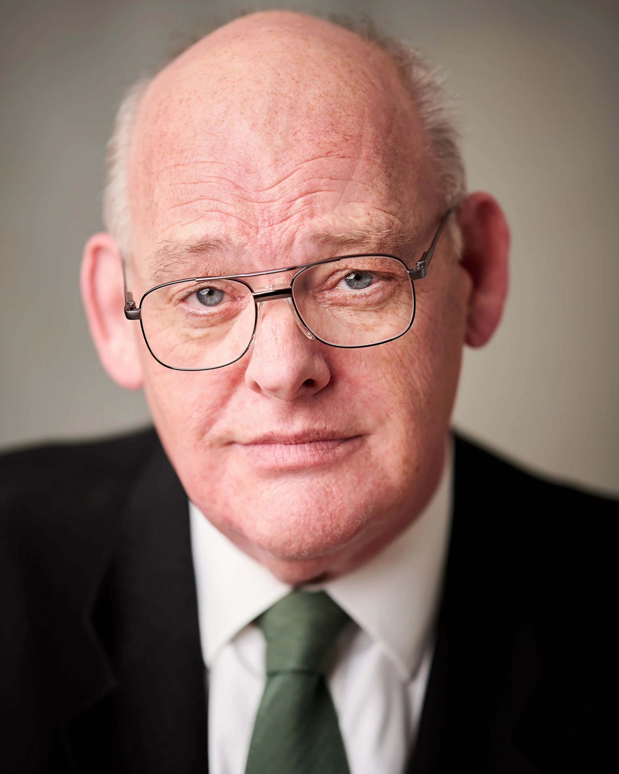 Close-up of an older man with glasses, receding hairline, and wrinkles on his forehead, dressed in a suit, white shirt, and tie.