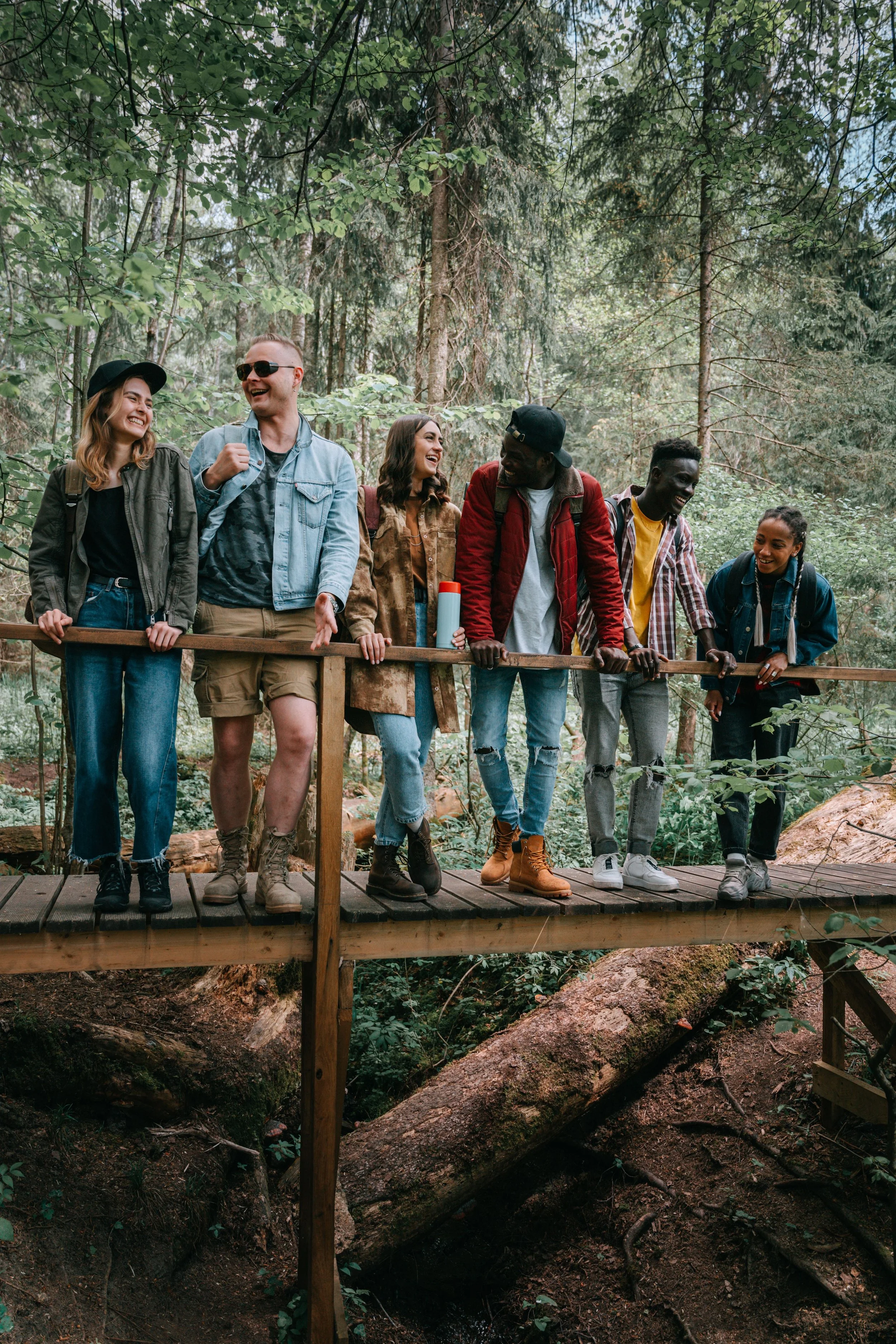 A group of six diverse young friends standing on a wooden bridge in a forest, smiling and laughing together.