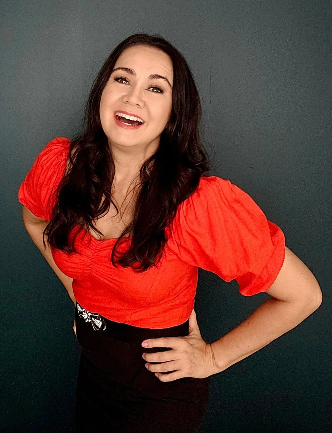 Mid-shot photo of woman with dark long hair, red blouse and black skirt, smiling with a big open smile, like mid laugh.
