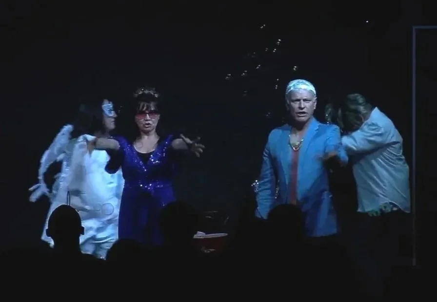 Two actors at the front of the stage singing and wearing sparkly costumes. There are bubbles around them and two dancers dressed in white behind them