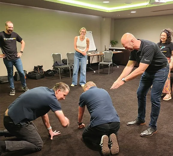 An improv workshop at an executive leadership conference. Three of the participants, who are male, are either on all fours or crouching over with diving hands. Other participants are standing around them laughing.