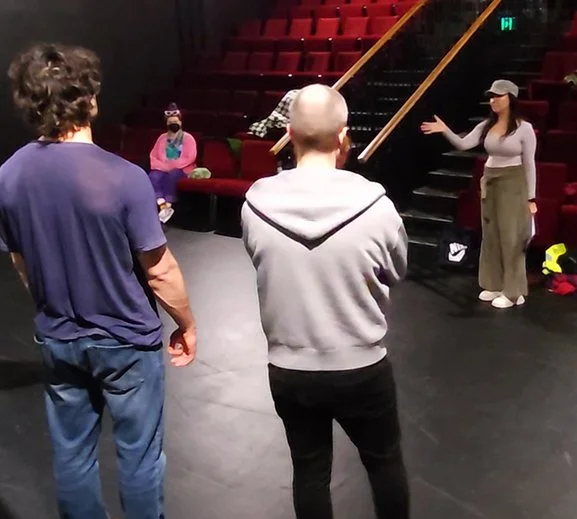 A woman wearing a cap is coaching performers on stage. There are red theatre seats behind her the rise up.