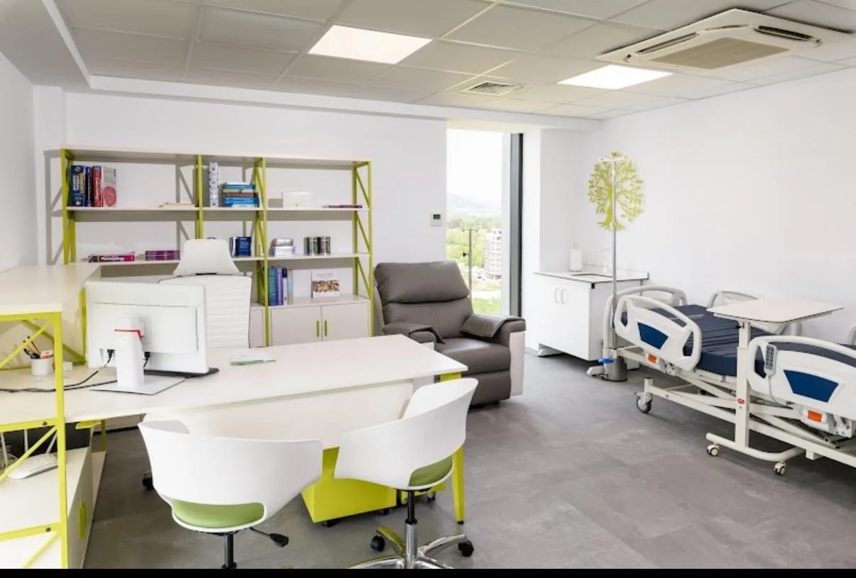 Ein Krankenzimmer mit einem Krankenhausbett, einem Sessel, Schreibtisch, Bücherregal, Computer und einem Fenster mit Blick nach draußen.