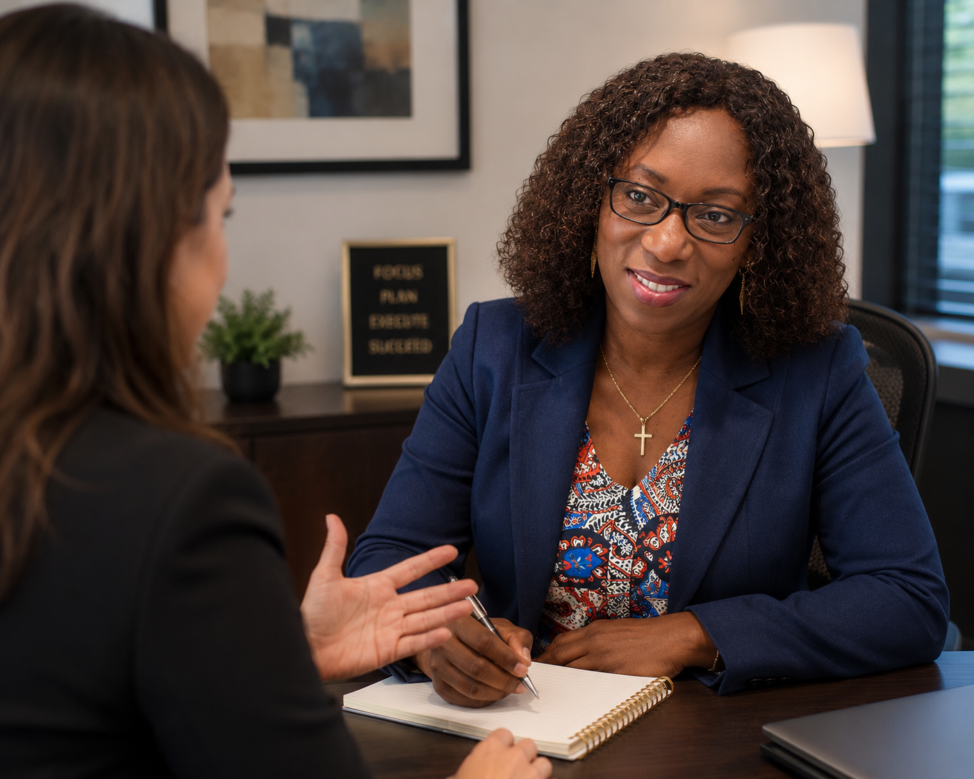 Two women having a conversation in an office, one is speaking and the other is listening with a notebook and pen in front of her.