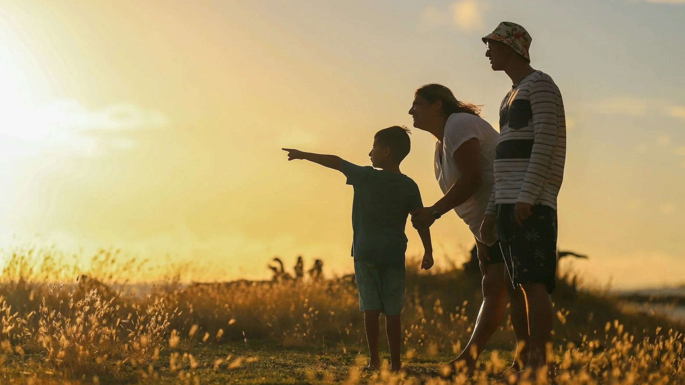 Four people, including children and adults, walk through a field at sunset, with one child pointing into the distance.