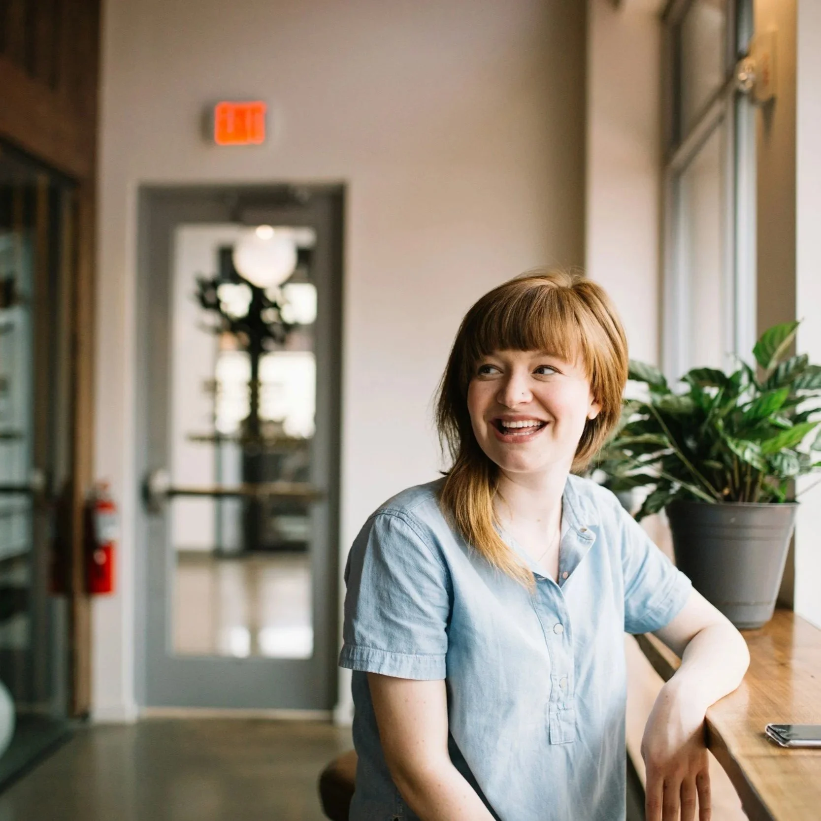A young woman with reddish-brown hair and a nose ring sitting at a wooden counter in a well-lit room, smiling and looking to her left, with a large potted plant beside her and a door and exit sign in the background.