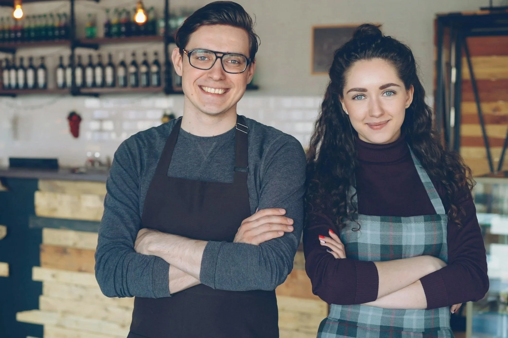 A young man and woman standing inside a coffee shop, smiling at the camera. The man is wearing glasses and a dark gray shirt with a black apron, and the woman has curly dark hair, blue eyes, and is wearing a dark long-sleeve shirt with an apron.