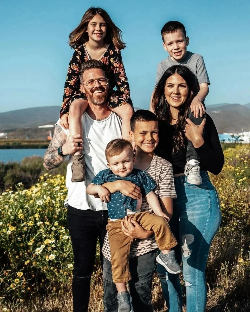 A family of seven posing outdoors in a sunny field with mountains in the background. The children are sitting or on the parents' shoulders, smiling and dressed casually.