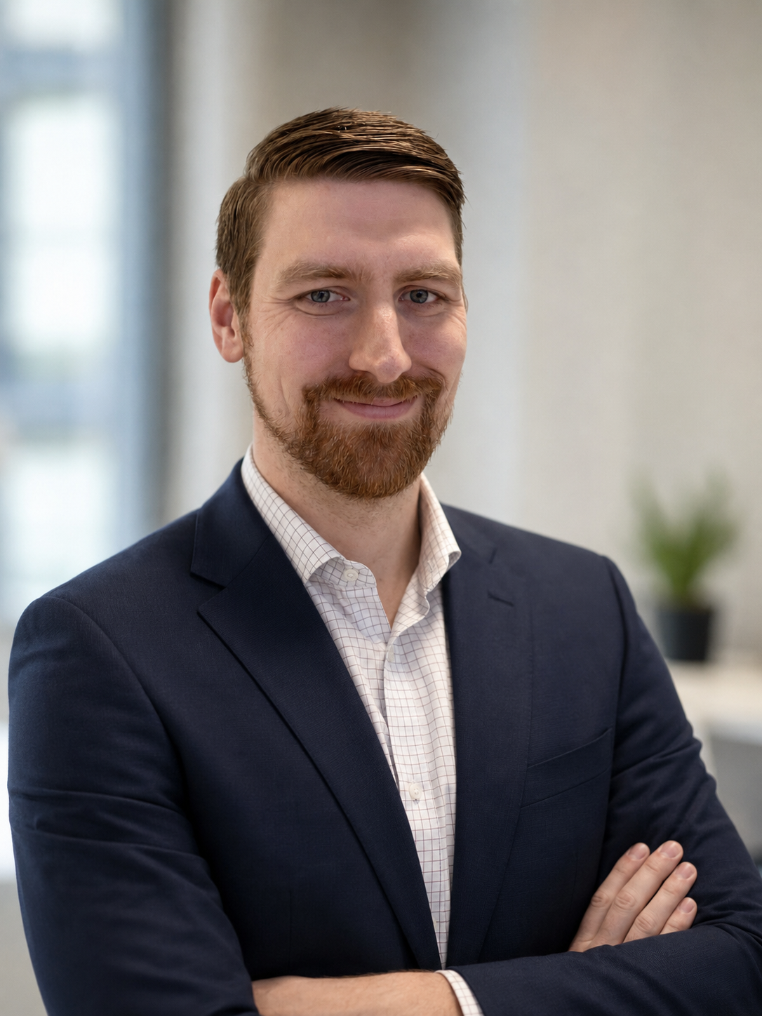 A professional man with red hair and a beard, dressed in a dark blazer and checkered shirt, posing with arms crossed in an office setting with blurred background and a potted plant.