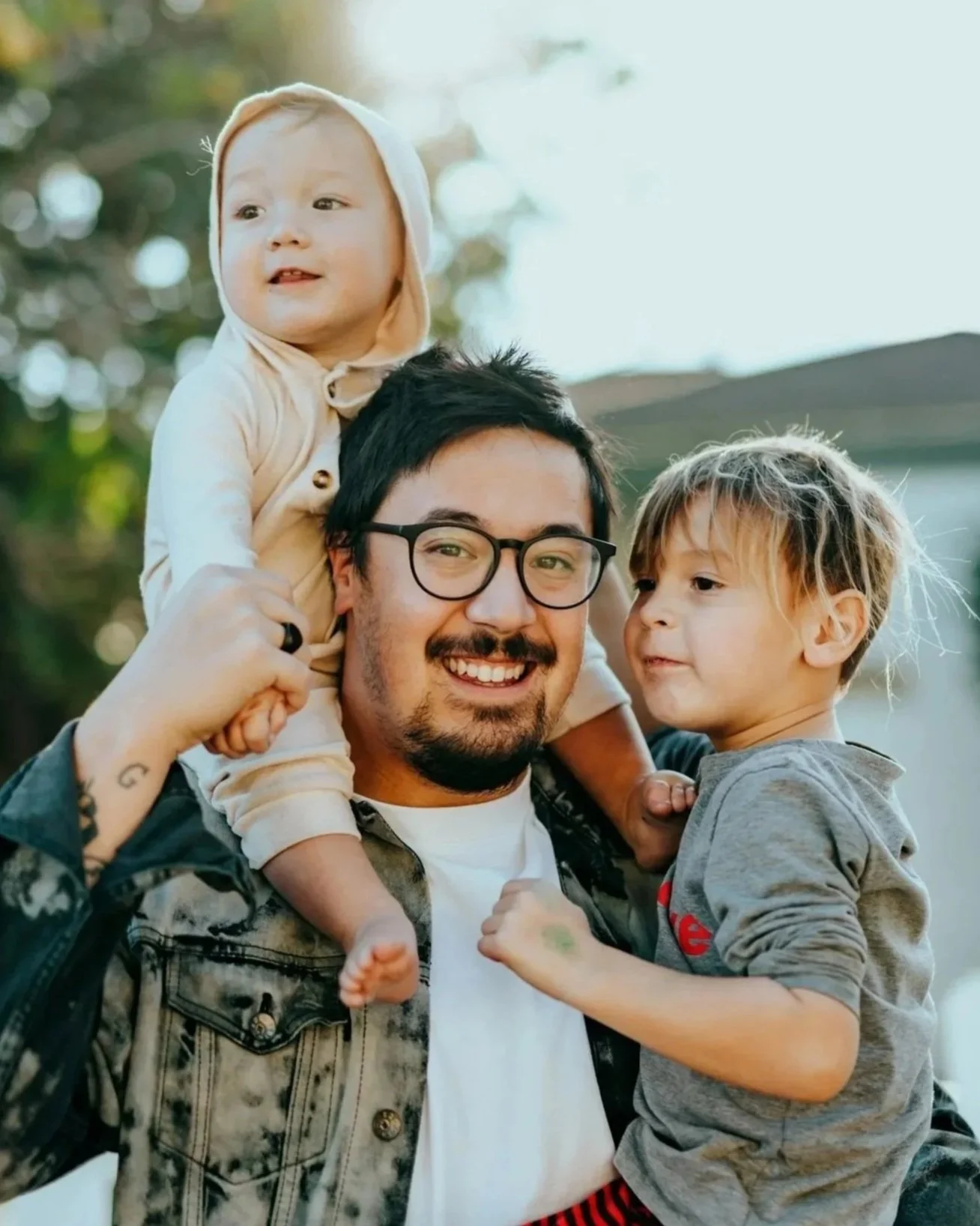A man with glasses and a beard smiling as two children, a boy and a baby girl, sit on his shoulders outdoors during daytime with trees and sunlight in the background.