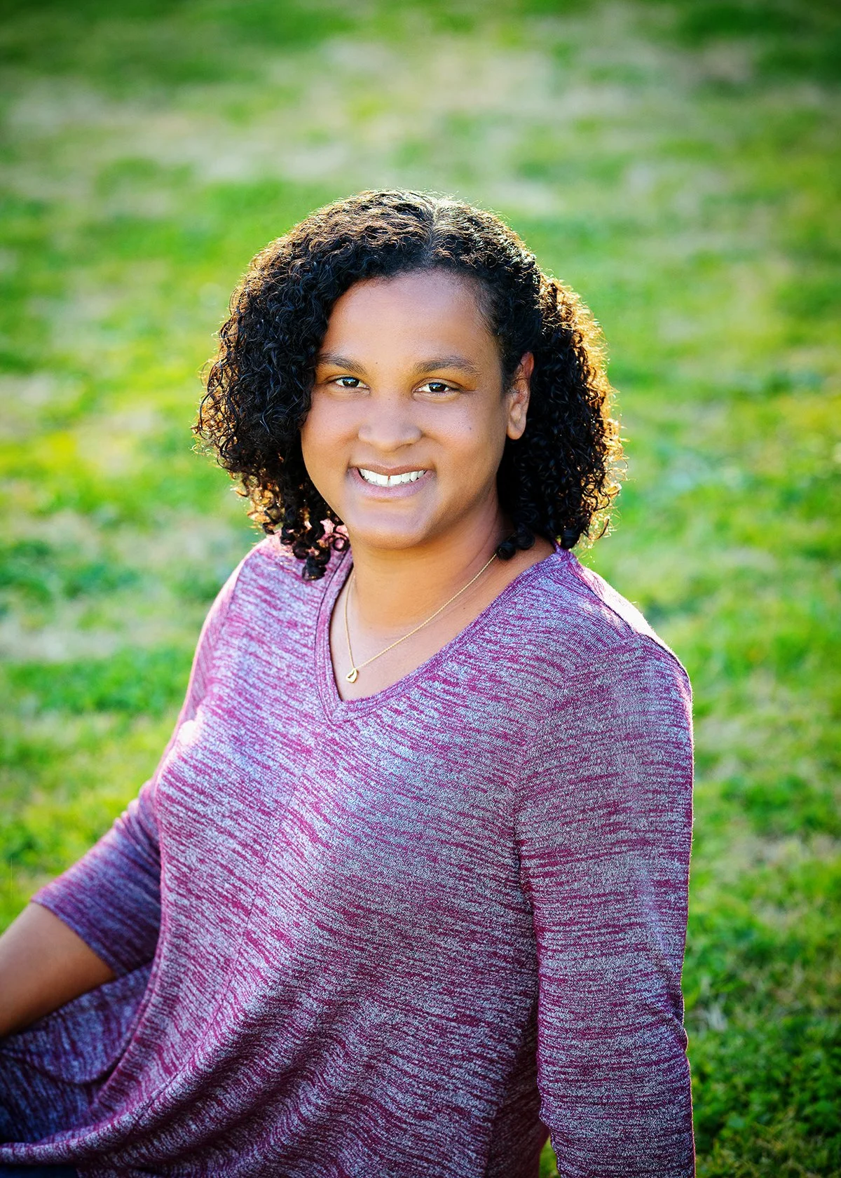 A woman with curly dark hair wearing a maroon long sleeve top and a delicate gold necklace, sitting outdoors on grass in sunlight, smiling.