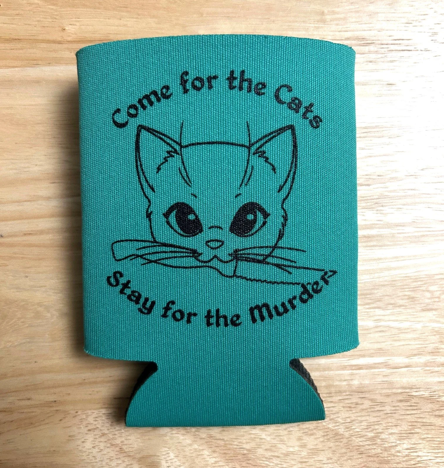 A teal-colored foam can cooler with a black embroidered illustration of a cat's face and text around it that says 'Come for the Cats, stay for the Murder'.