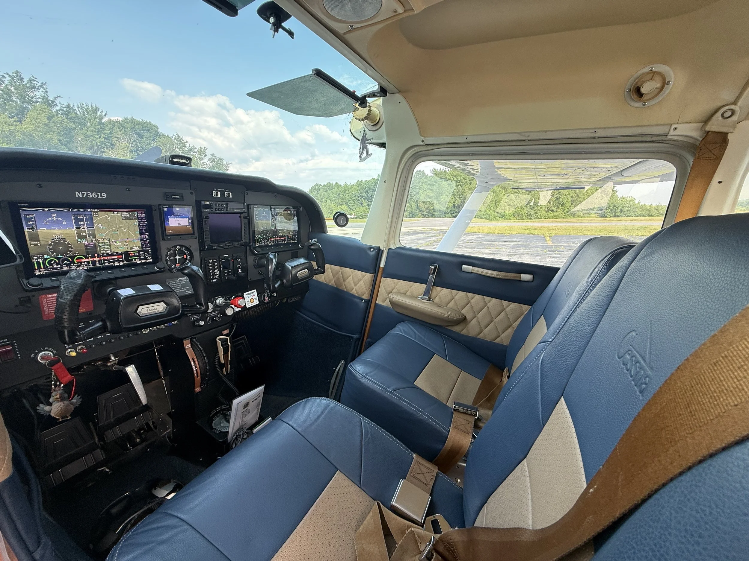 Cockpit interior of an airplane showing the instrument panel, seats, and a large window revealing a view of trees and clouds outside.
