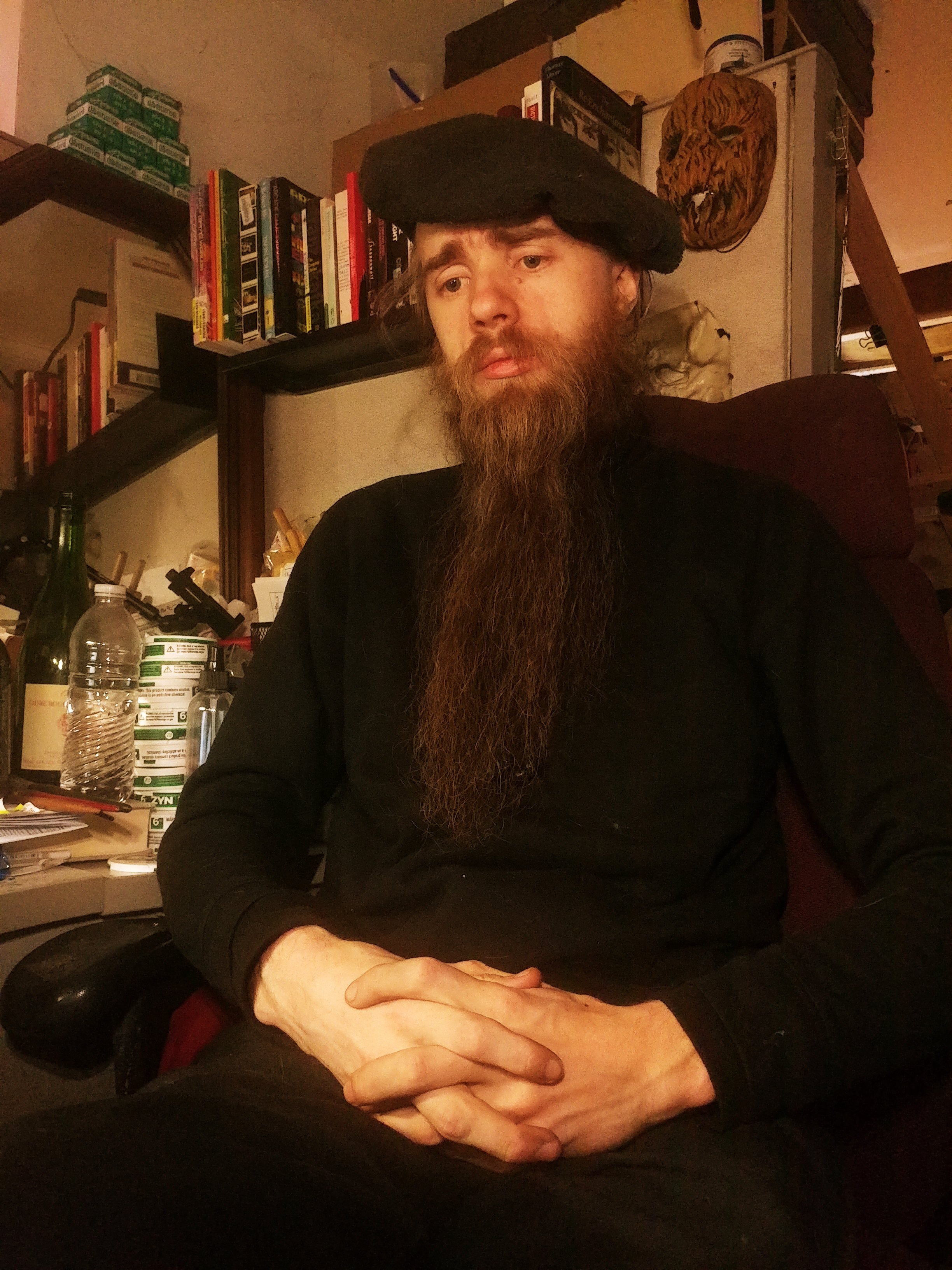 A man with a long beard and mustache wearing a black hat sits at a table with his hands clasped. Behind him are shelves with books and various items, including a framed picture of a tiger's face, some canned goods, and bottles.