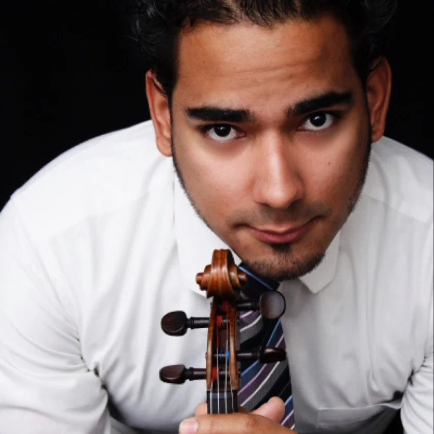 Close-up of a man in a white shirt and striped tie holding a violin, looking directly at the camera against a black background.