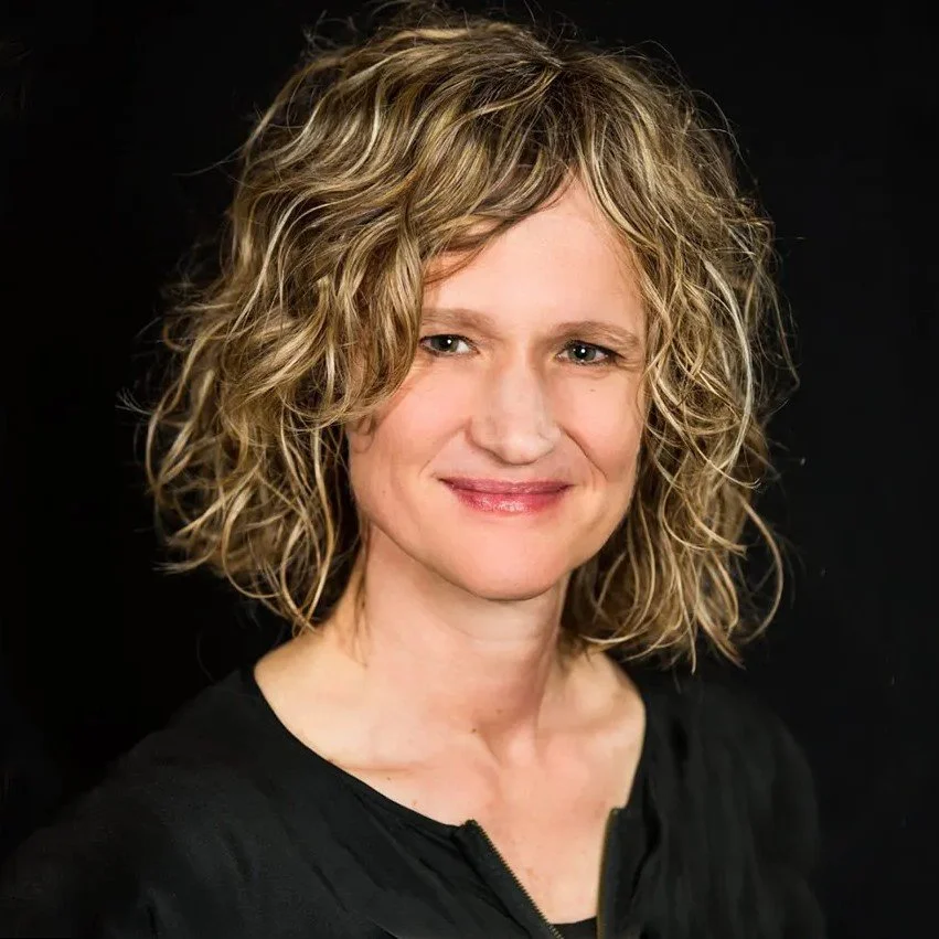 A woman with curly blond hair smiling against a black background.