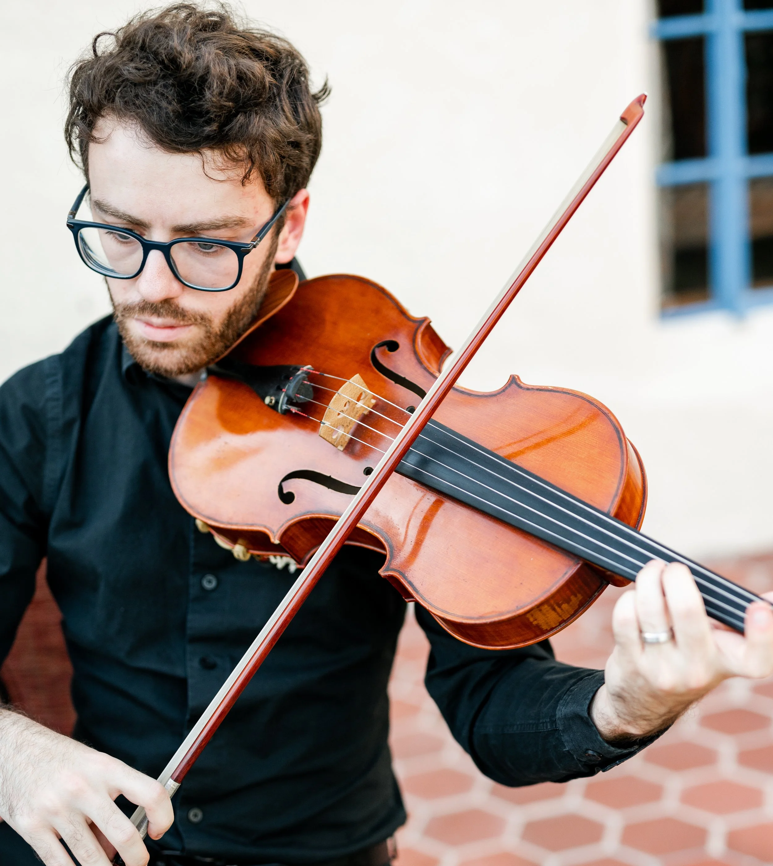 A man with curly brown hair and glasses playing a violin indoors. He is wearing a black button-down shirt and is focused on his playing, with a blurred background showing part of a blue shelf and a tiled floor.