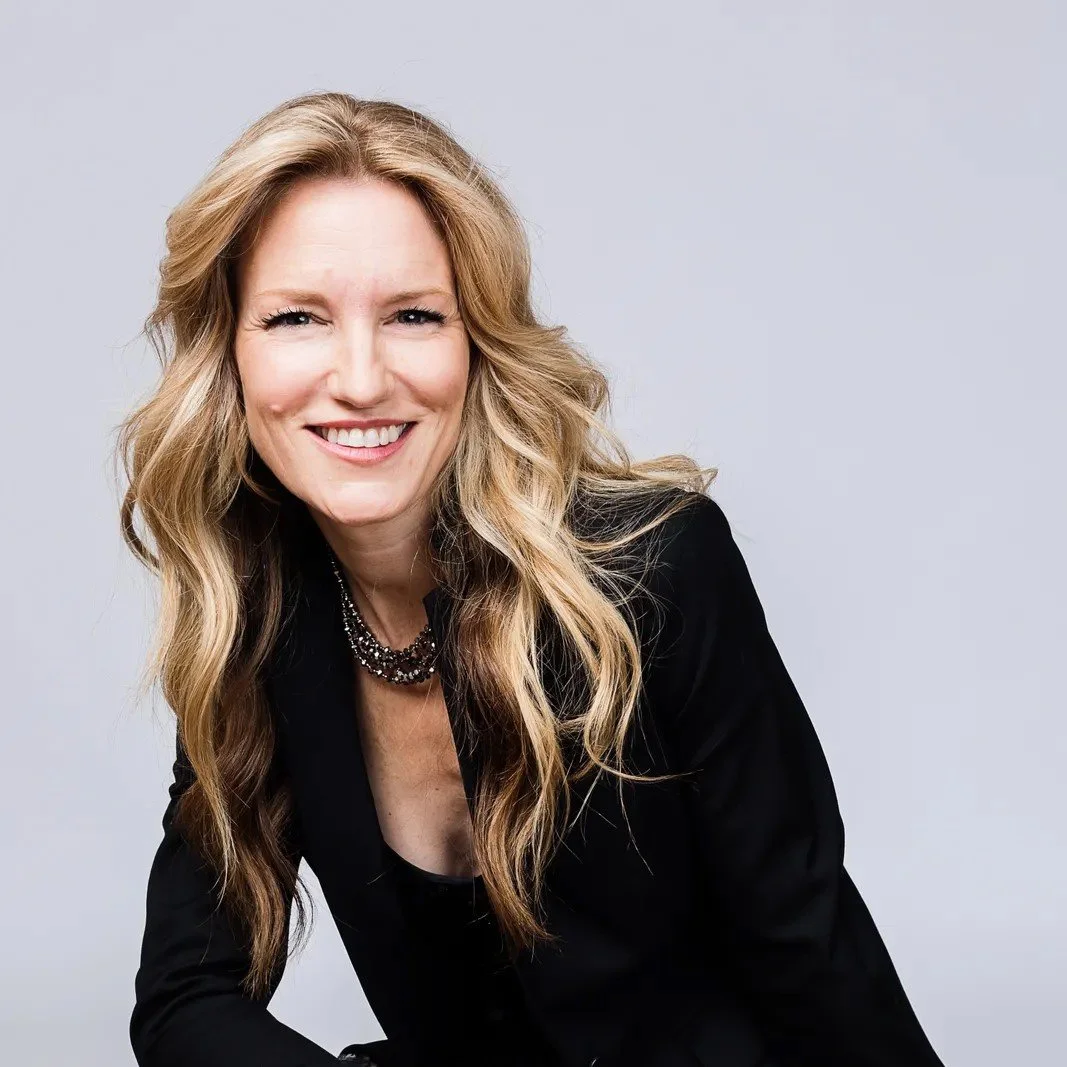 Portrait of a woman with long wavy blonde hair, smiling, wearing a black blazer and a layered necklace, against a plain light background.