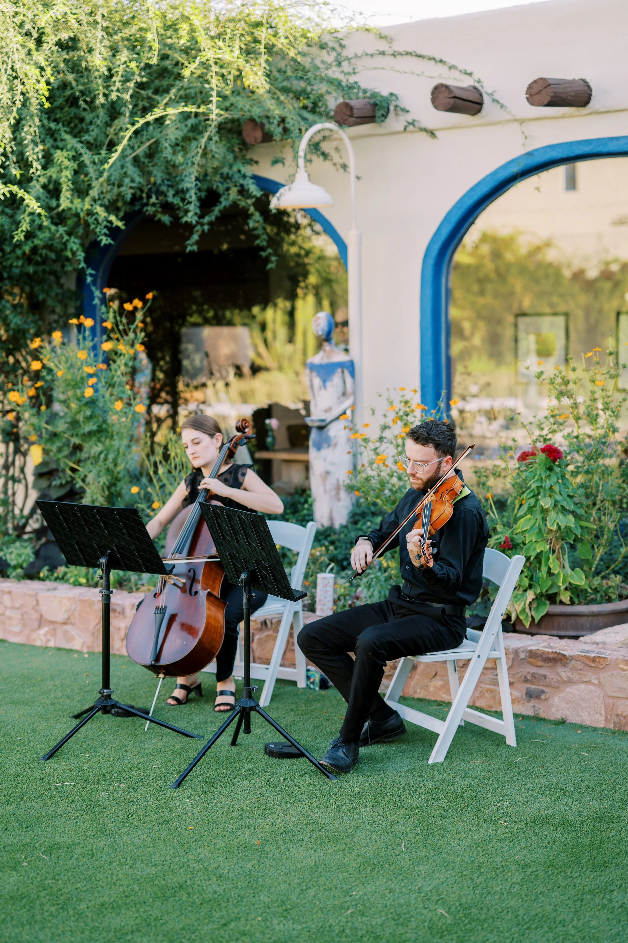 A duo of musicians playing string instruments outdoors, seated on white chairs in a garden with lush greenery and yellow flowers, under a white building with blue accents.